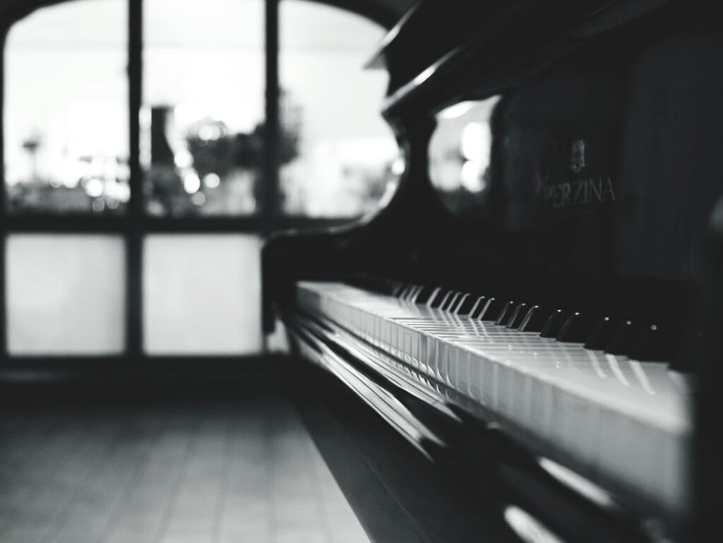 black and white artistic image of piano in a converted industrial space taken from one end of the keys, out of focus mezzanine window in background, city scene