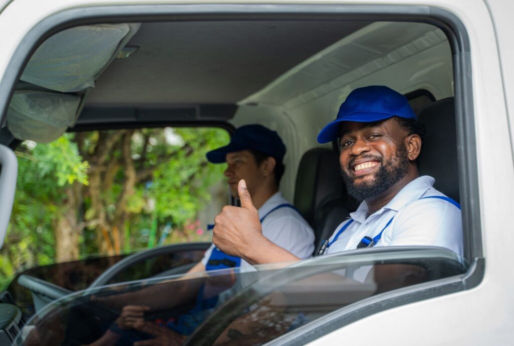 Two professional movers in the cab of a truck. Man in driver's seat smiles and gives thumbs up.