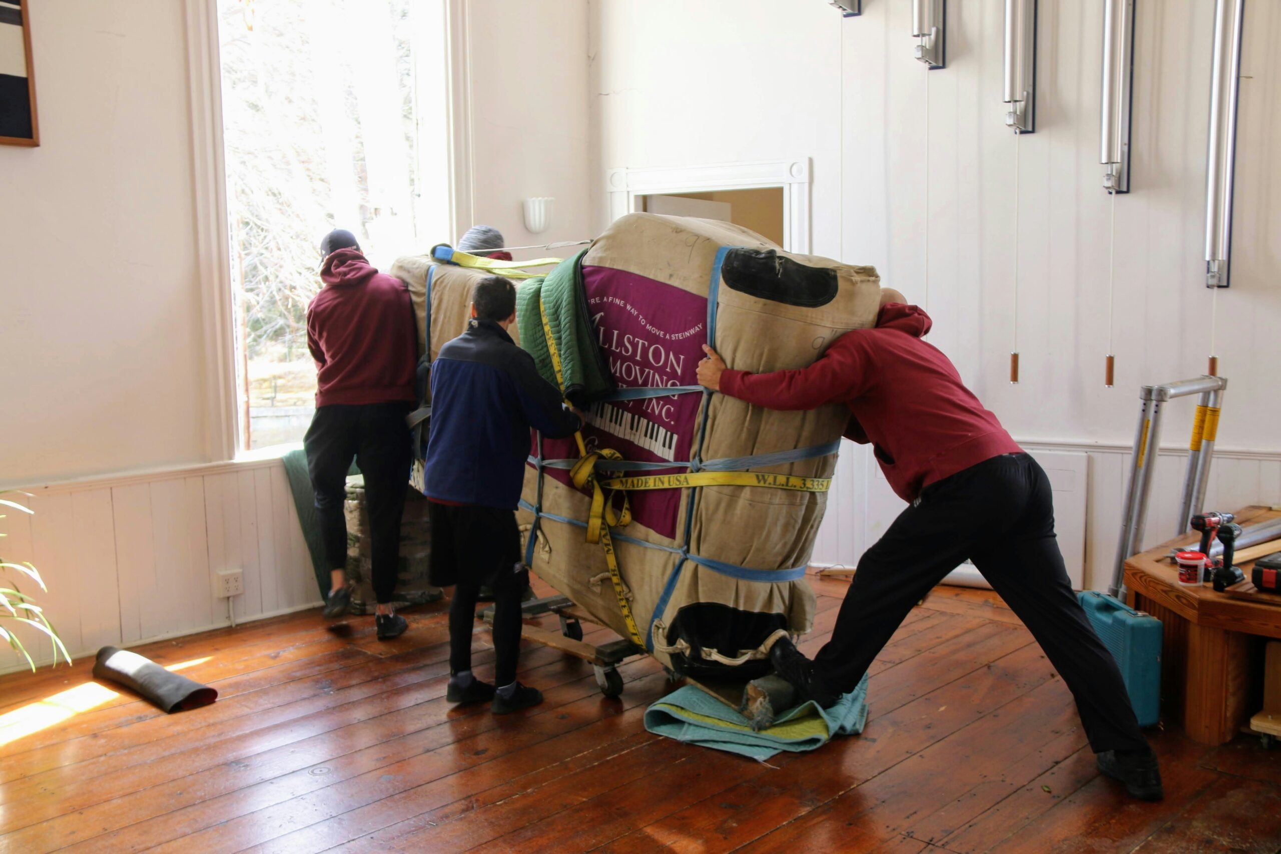 Two professional piano movers in navy shirts carefully maneuver a grand piano on a dolly through a tight historic Asheville home doorway, navigating scuffed floors and baseboards, illuminated by filtered sunlight through antique windows, emphasizing expert care in challenging moving conditions.